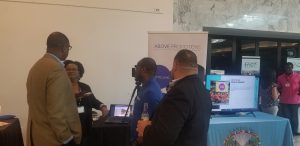 Above Promotions in Soaring Cities 100 Tables of Innovation, Four people stand and talk near a booth for a digital marketing agency, with banners, a monitor displaying information, and promotional materials at an indoor event.