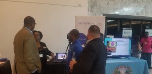 Above Promotions in Soaring Cities 100 Tables of Innovation, Four people stand and talk near a booth for a digital marketing agency, with banners, a monitor displaying information, and promotional materials at an indoor event.