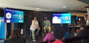 Above Promotions in Synapse Summit Tampa 2023, Ebony and Ronald Vaz sit on stage at the Black Tech Talent & Innovator Gathering, joined by digital marketing agency professionals in the audience, with presentation slides displayed on two screens.