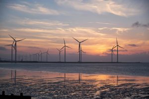 Silhouetted wind turbines stand in a line along a coastal area at sunset, with a cloudy sky and reflections on the wet sand below.