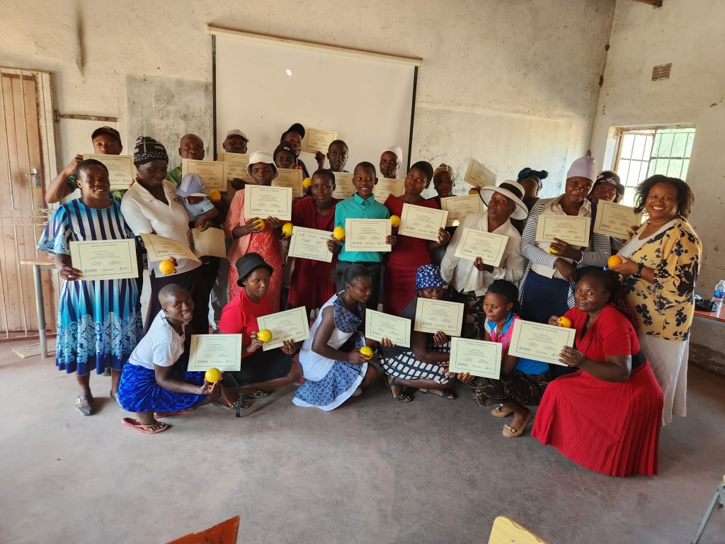 A group of people proudly display certificates and oranges, posing indoors in a casual setting.