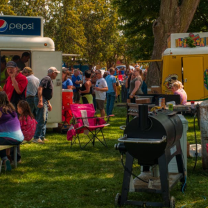 Outdoor food festival with people gathered around a Pepsi truck and another vendor. Hot dog stand visible, with chairs and a grill in the foreground. Trees and grass surround the event area.