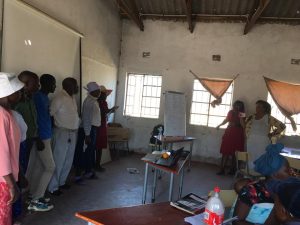 Above Promotions in Zimbabwe; A group of people stand near a whiteboard in a classroom with bare walls and windows, as two women from a digital marketing agency appear to be leading a discussion.