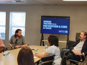 Four people sit around a conference table with water bottles, facing a screen displaying "Working together for the prevention & cure of cancer. Moffitt Cancer Center," during a digital marketing agency strategy session.