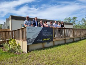 Eight people stand on a wooden deck in front of a building with a sign reading "A NEW BEGINNING STARTS HERE — SPEROS FL," home to a life sciences park and a digital marketing agency.