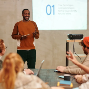 A person in a brown sweater presents to a group, holding a tablet. A slide projector displays "01" and text about brand identity. Attendees are seated around a table with laptops.