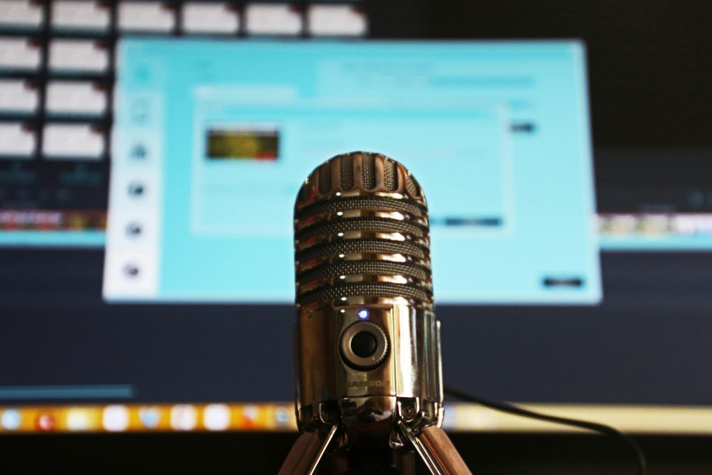 Silver microphone in focus with a blurred computer screen in the background displaying an editing software interface. Silver microphone in focus with a blurred computer screen in the background displaying an editing software interface.