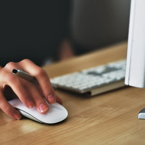 A hand using a computer mouse on a wooden desk, with a keyboard and monitor in the background.