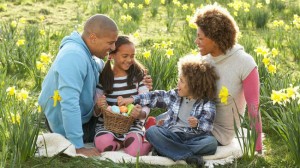 A family of four enjoying an Easter egg hunt in a field of daffodils. The parents and two children are smiling and examining colorful eggs in a basket, capturing the moment for their social media followers.