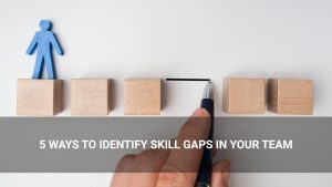 A hand draws a line between wooden blocks with a blue human figure on one block, illustrating how to identify skills gaps in your team; text overlay reads, "5 ways to identify skill gaps in your team.