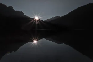 The sun sets behind a mountain range, casting a bright starburst and its reflection on a calm body of water.