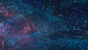Abstract image of a colorful starry sky with blue, pink, and white speckled patterns resembling cosmic dust against a dark background.
