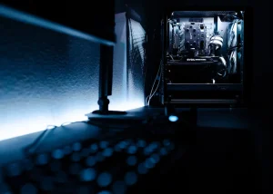 A dimly lit room with a glowing computer case displaying internal components. A keyboard and monitor are partially visible in the foreground.