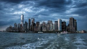 View of New York City skyline from the water at dusk, with illuminated skyscrapers under a cloudy sky and a boat on the river.