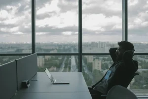 Person sitting at a desk with a laptop, looking outside a large window showing a cityscape under a cloudy sky.